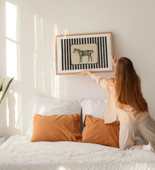 A woman in a bedroom adjusts a framed art print hanging above a bed. The print features a black and white striped background with a central beige rectangle containing a black horse illustration.