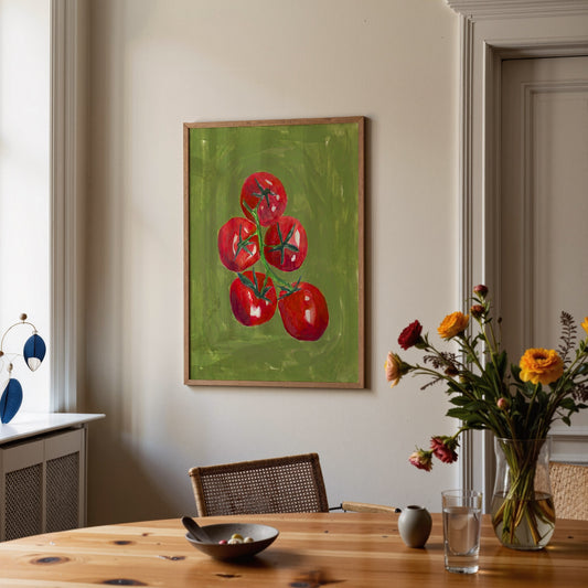 A framed painting of red tomatoes on a green background hanging on a beige wall in a dining room setting with a wooden table and flowers.