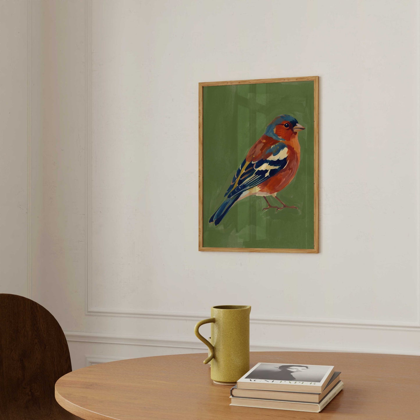 A framed art print of a bluebird on a green background, hanging on a white wall above a wooden table with a yellow-green mug and a stack of books.