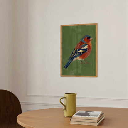 A framed art print of a bluebird on a green background, hanging on a white wall above a wooden table with a yellow-green mug and a stack of books.