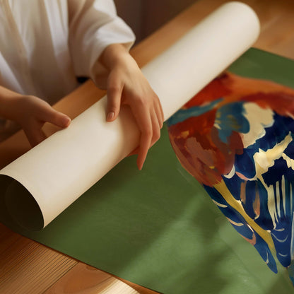 A person unrolls a large sheet of green paper or mat on a wooden table, with part of a colorful painted object visible underneath.