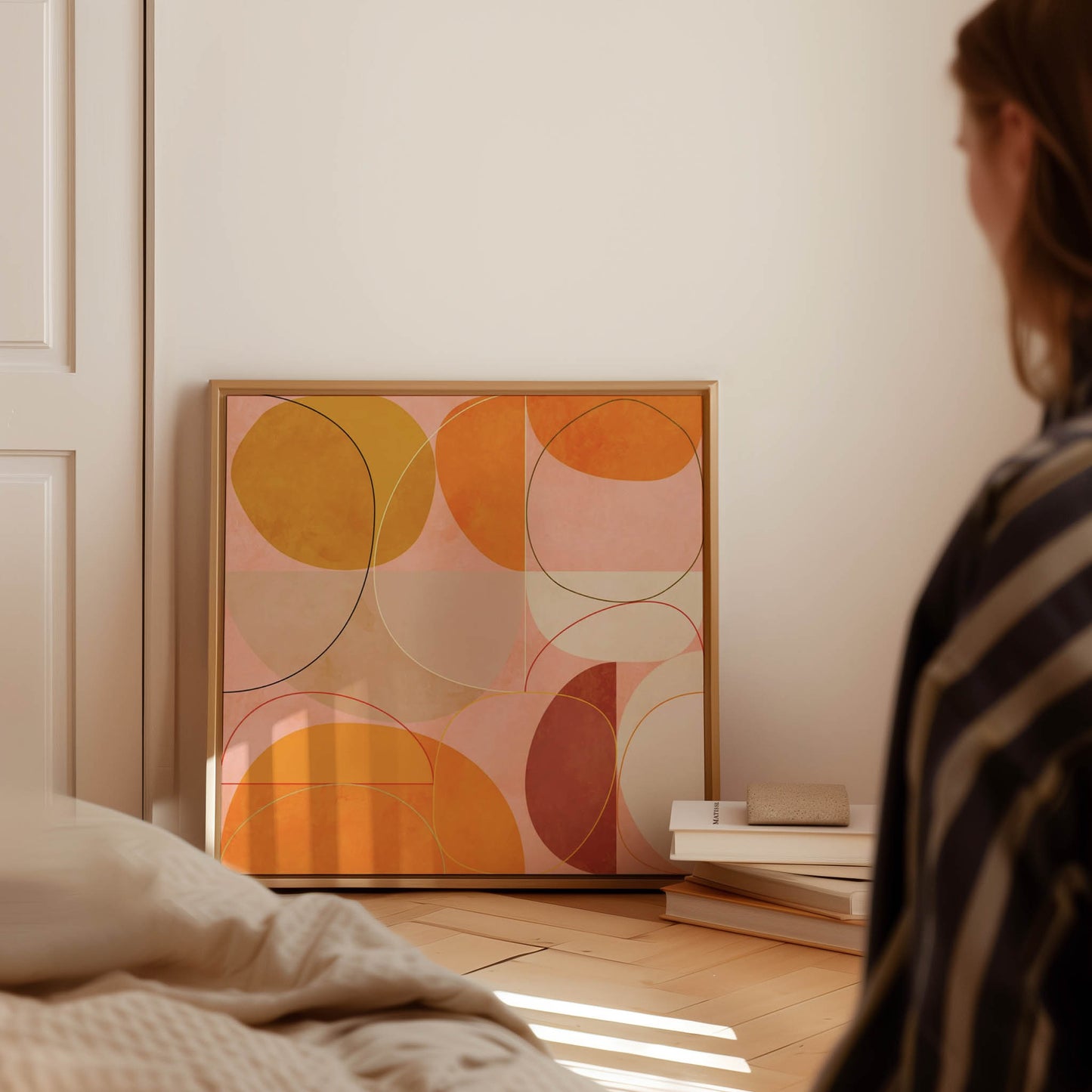A framed abstract art print with overlapping circles in orange, yellow, and pink tones, leaning against a wall in a softly lit bedroom. A person is partially visible in the foreground, and a bed and small stack of books are in the room.
