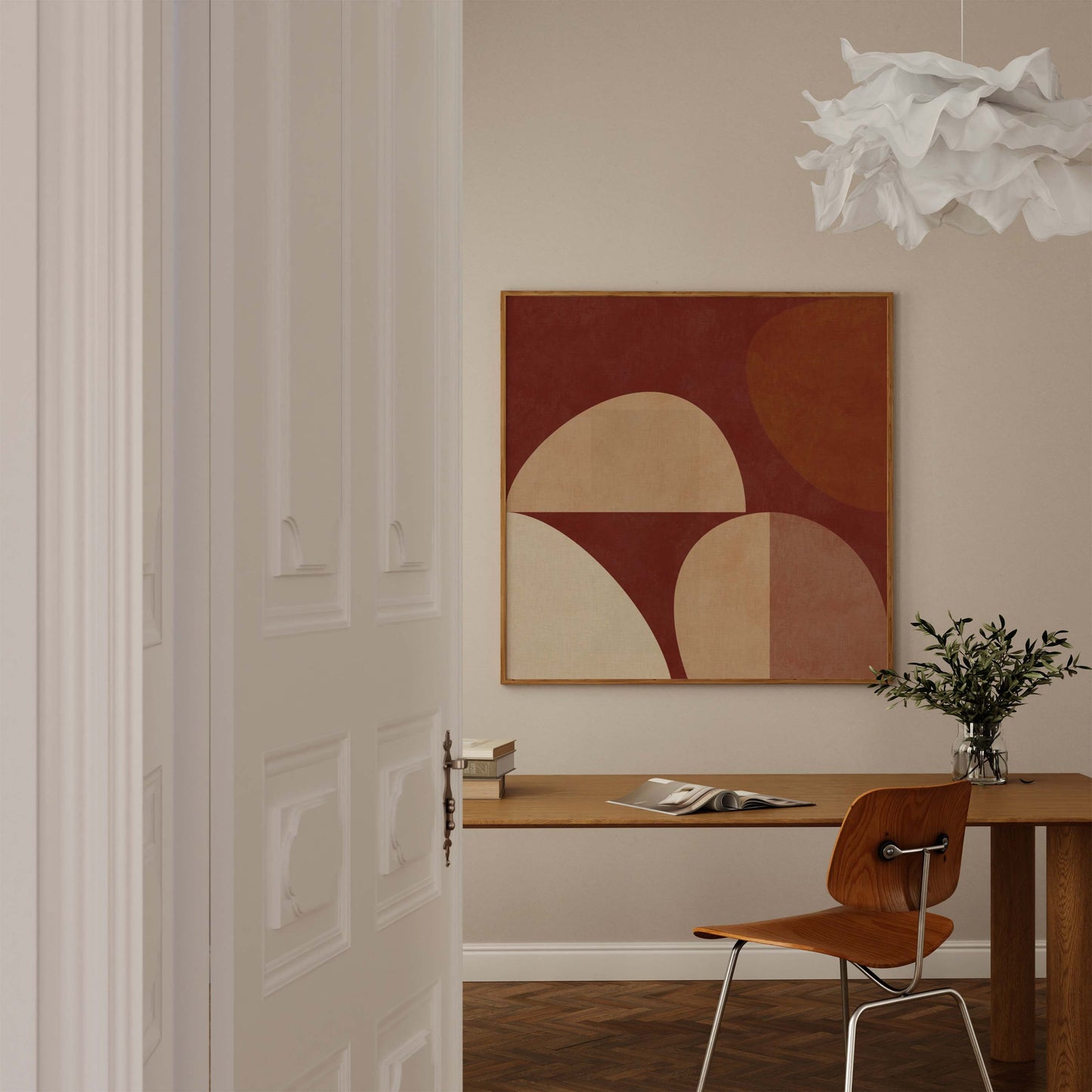 A framed abstract geometric wall art print hangs above a wooden desk in a warm, modern room with a chair, vase of greenery, and a white paper lantern light fixture.