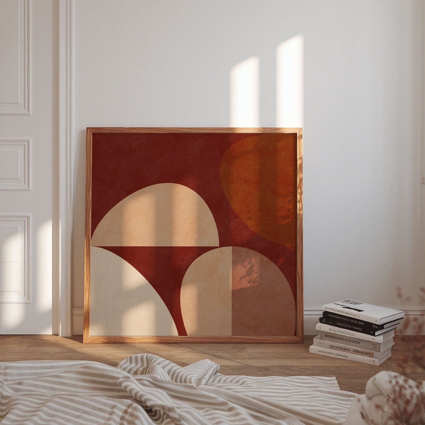 Framed abstract wall art leaning against a white wall in a sunlit room, with a stack of books and a striped blanket in the foreground.