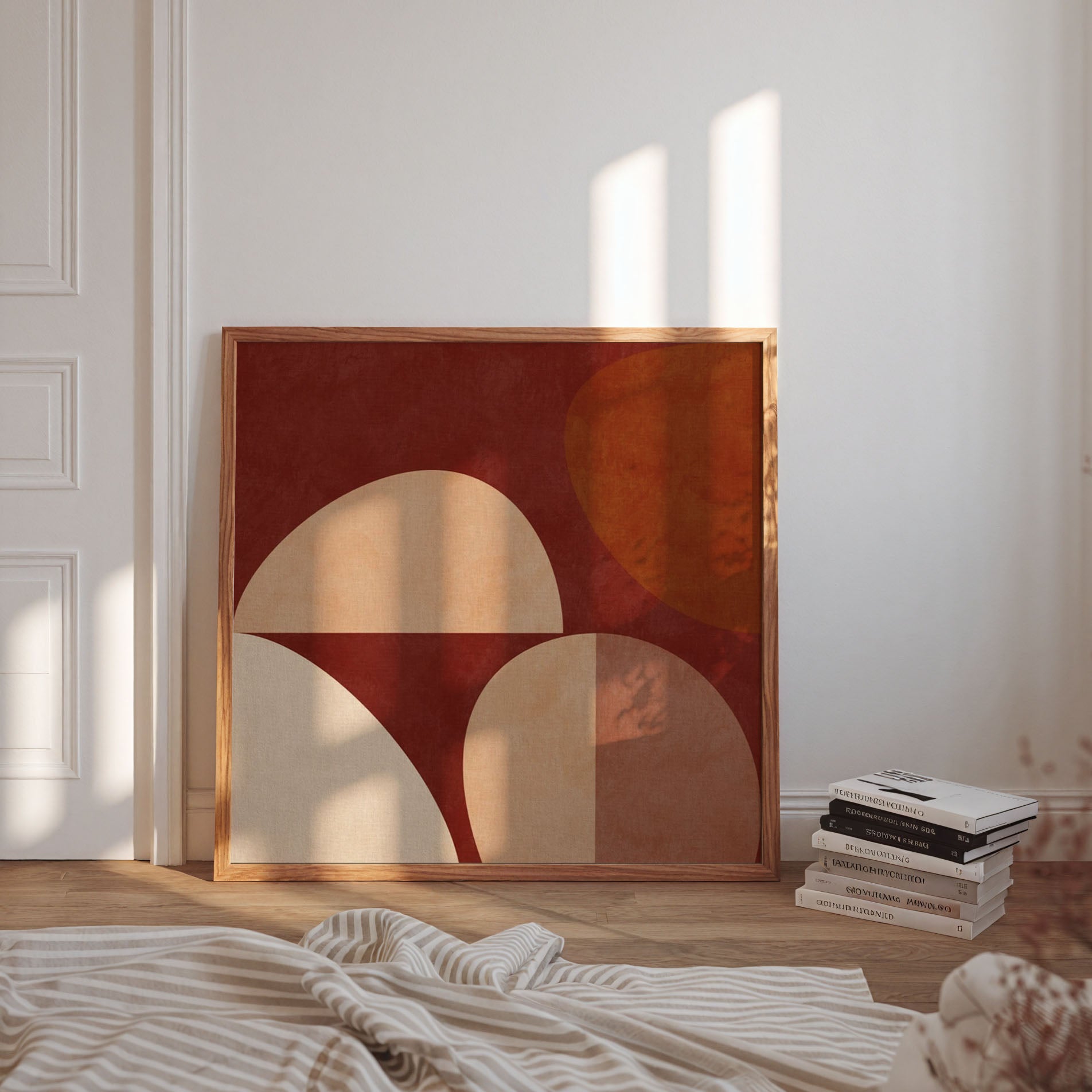 Framed abstract wall art leaning against a white wall in a sunlit room, with a stack of books and a striped blanket in the foreground.