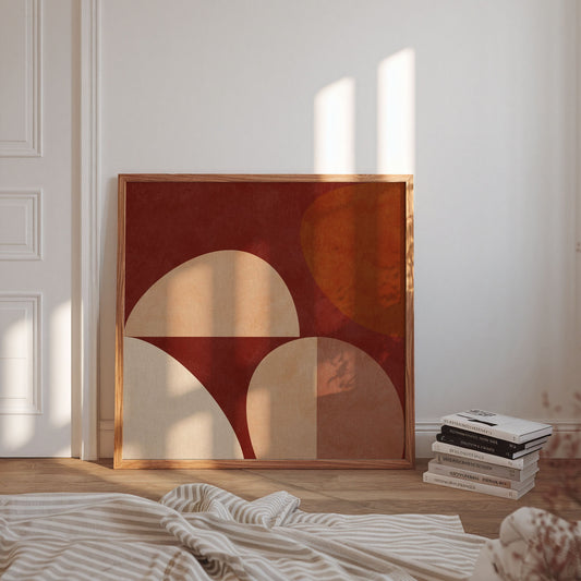Framed abstract wall art leaning against a white wall in a sunlit room, with a stack of books and a striped blanket in the foreground.