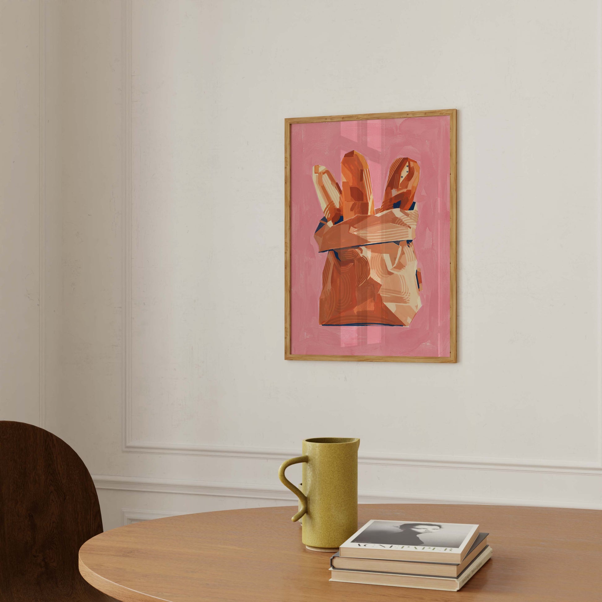 A framed art print featuring a pink background and a hand holding bread loaves, hanging on a white wall above a wooden table with a green mug and books.