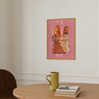 A framed art print featuring a pink background and a hand holding bread loaves, hanging on a white wall above a wooden table with a green mug and books.