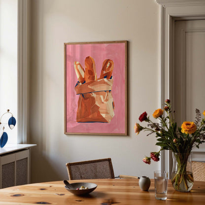 A framed art print depicting a pink background with a brown paper bag containing baguettes, hanging on a beige wall in a dining room setting with a wooden table, chairs, and a vase of flowers.