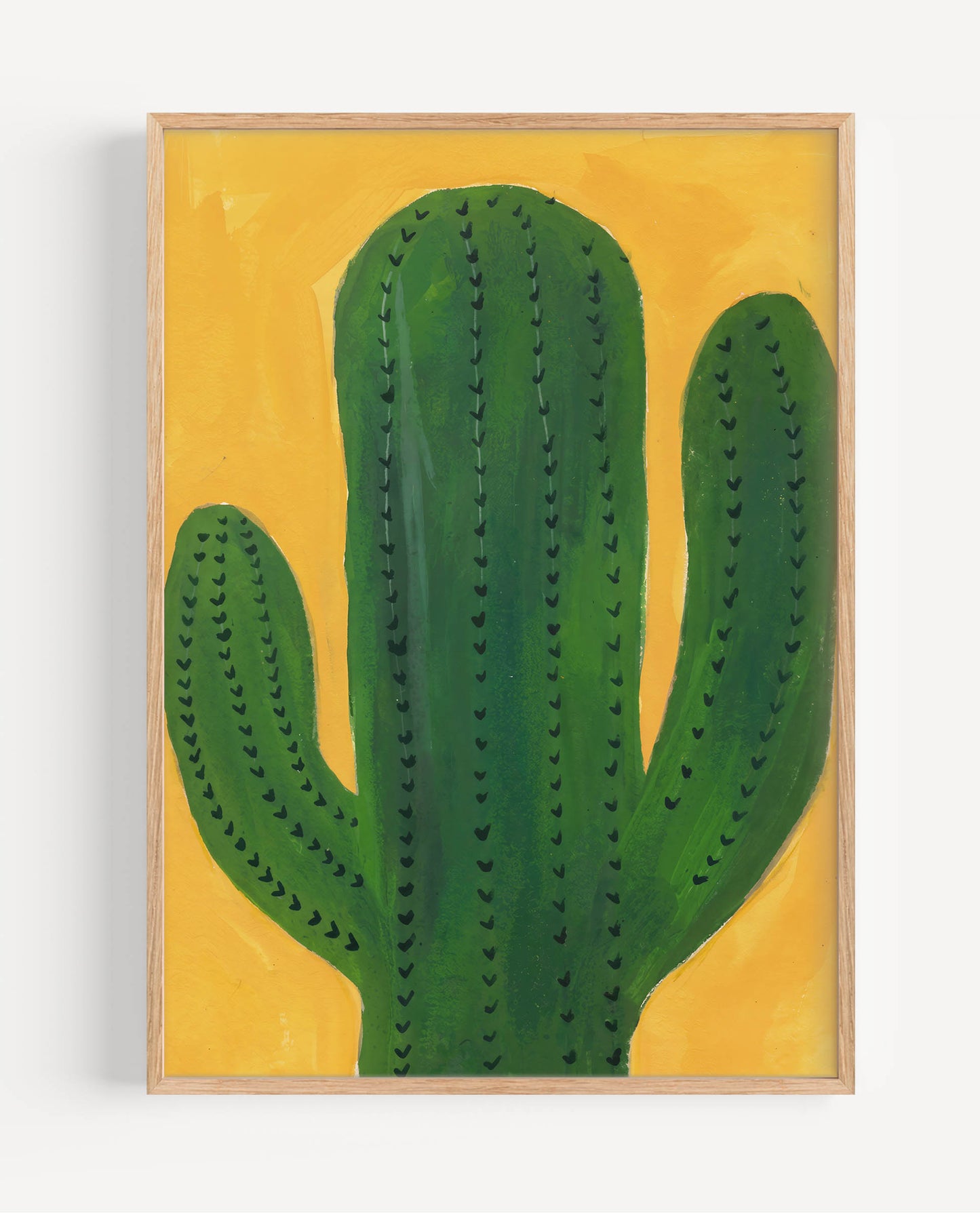A framed art print featuring a green cactus painted on a textured yellow background, set in a light natural wood frame against a white wall.