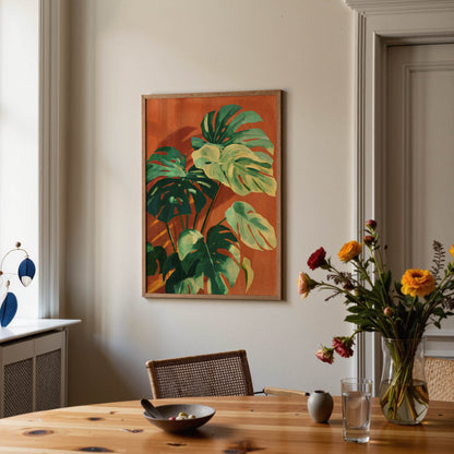 A framed botanical art print featuring green monstera leaves on an orange background, hanging on a beige wall in a styled dining room setting with a wooden table, flowers, and a chair.