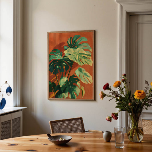 A framed botanical art print featuring green monstera leaves on an orange background, hanging on a beige wall in a styled dining room setting with a wooden table, flowers, and a chair.