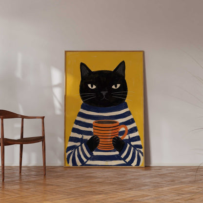 A framed art print depicting a black cat in a navy and cream striped sweater holding an orange mug, set against a yellow background, displayed on a wall in a room with a wooden chair and parquet floor.