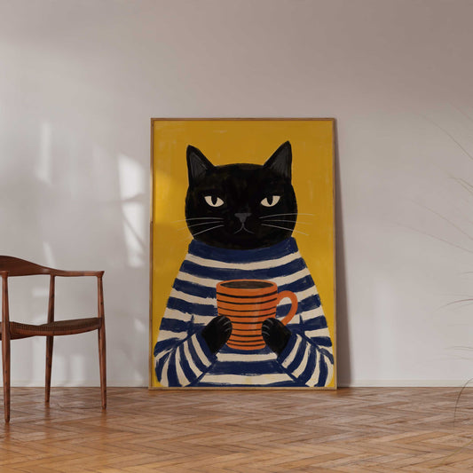 A framed art print depicting a black cat in a navy and cream striped sweater holding an orange mug, set against a yellow background, displayed on a wall in a room with a wooden chair and parquet floor.