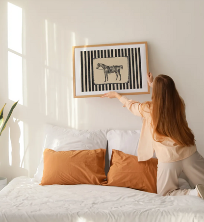A woman in a bedroom adjusts a framed art print hanging above a bed. The print features a black and white striped background with a central beige rectangle containing a black horse illustration.