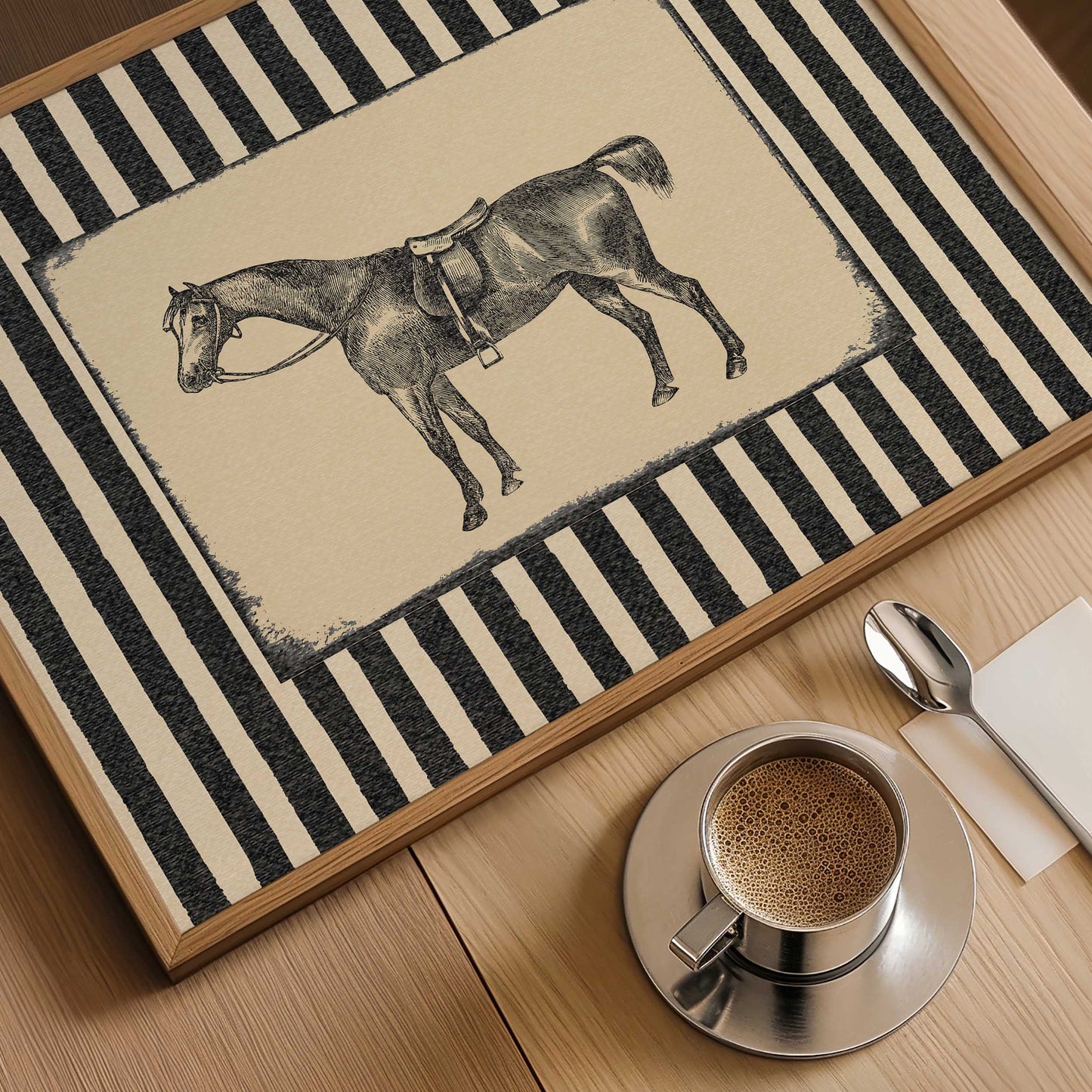 A rectangular framed rug or mat with a black-and-cream striped border and a central illustration of a horse wearing a harness, shown on a wooden table with a coffee cup, spoon, and napkin nearby.