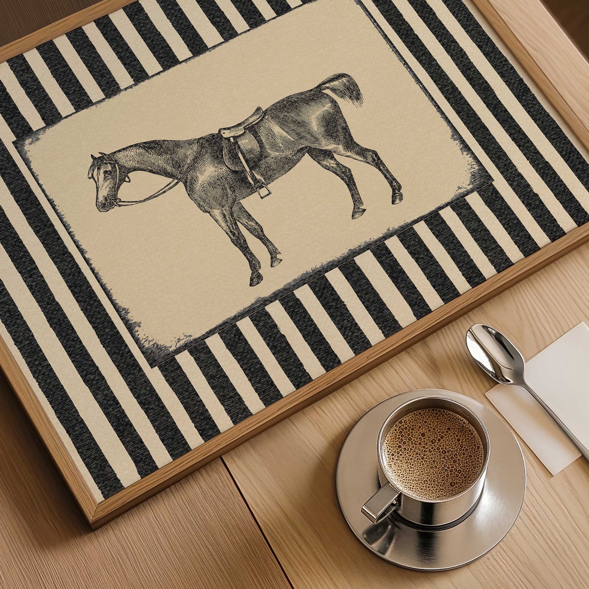 A rectangular framed rug or mat with a black-and-cream striped border and a central illustration of a horse wearing a harness, shown on a wooden table with a coffee cup, spoon, and napkin nearby.
