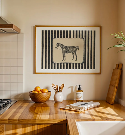 Framed wall art featuring a black-and-cream horse illustration on a striped background, displayed above a wooden kitchen counter with bowls, utensils, and a soap dispenser in a warm beige kitchen setting.