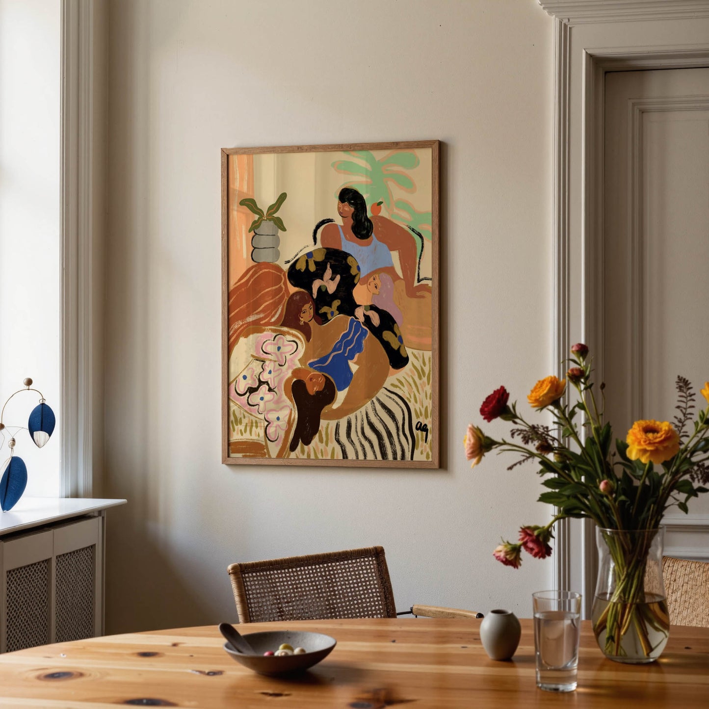A framed art print depicting an abstract, colorful scene of figures in a zebra-striped landscape, hanging on a beige wall in a modern dining room setting with a wooden table and flowers.