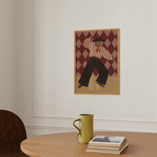 A framed art print featuring a stylized figure in a striped shirt and dark pants against a harlequin diamond background, hanging on a white wall above a wooden table with a green mug and stacked books.