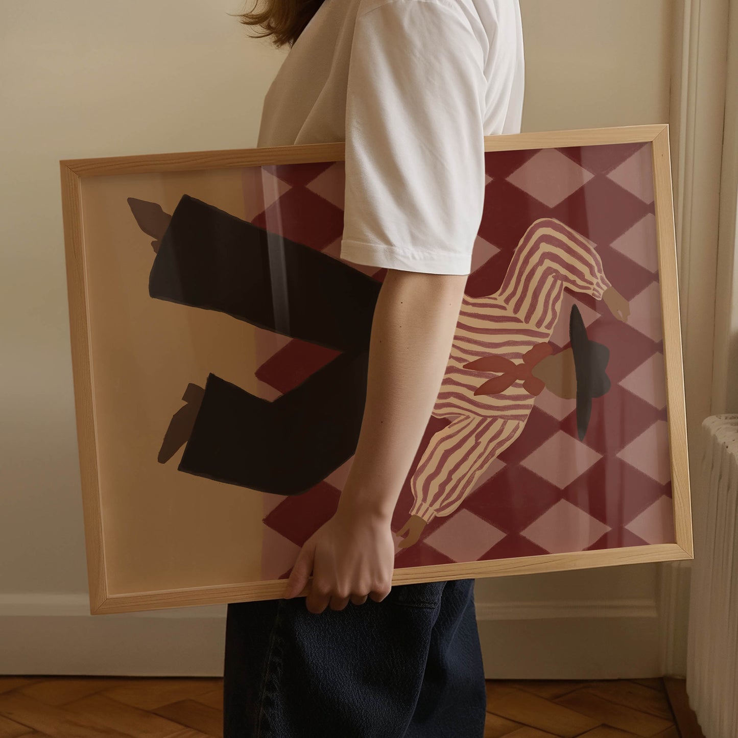 A person in a white t-shirt and dark jeans holds a large framed art print. The print features a stylized illustration of a zebra and black pants against a red and beige harlequin pattern, enclosed in a light wood frame.