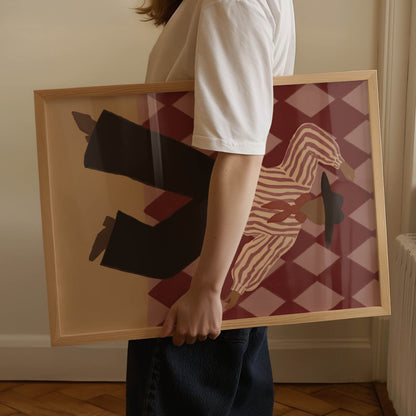 A person in a white t-shirt and dark jeans holds a large framed art print. The print features a stylized illustration of a zebra and black pants against a red and beige harlequin pattern, enclosed in a light wood frame.