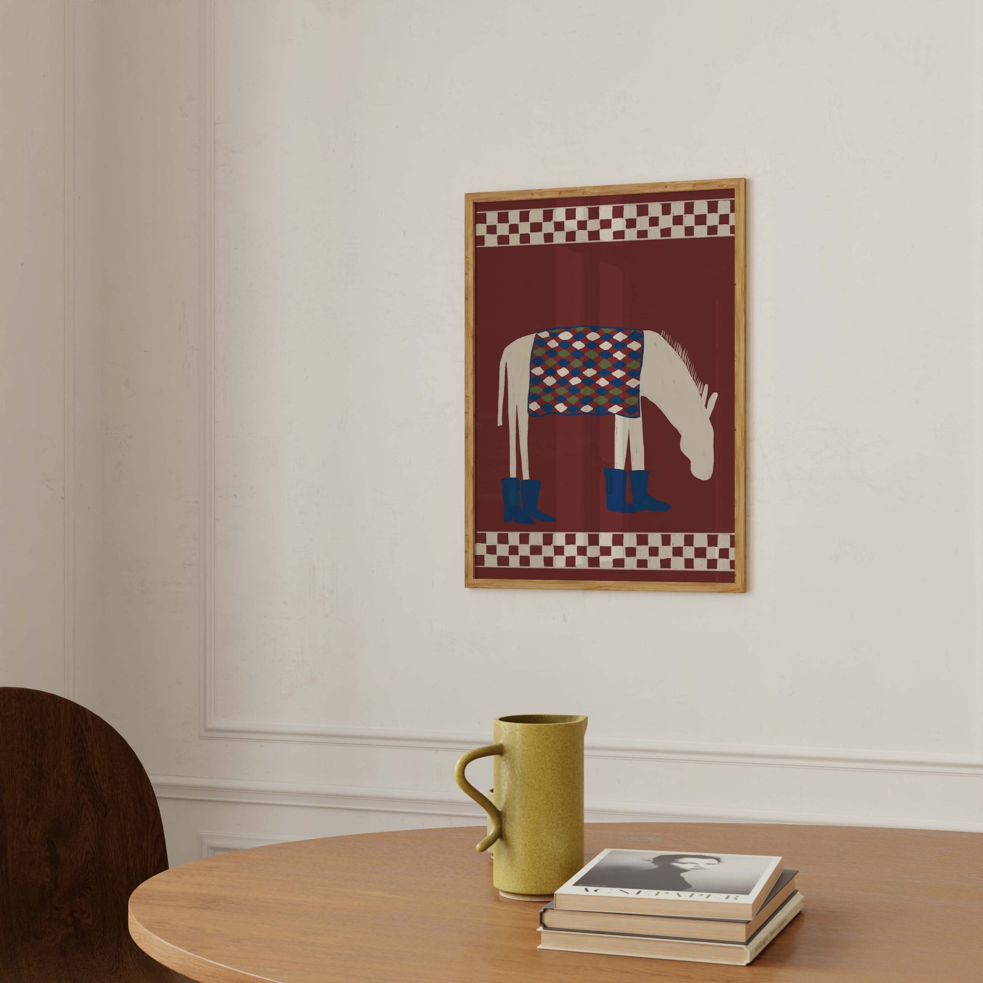 A framed art print featuring a stylized white horse with a blue patterned blanket and blue boots, hanging on a white wall above a wooden table with a yellow-green mug and books.