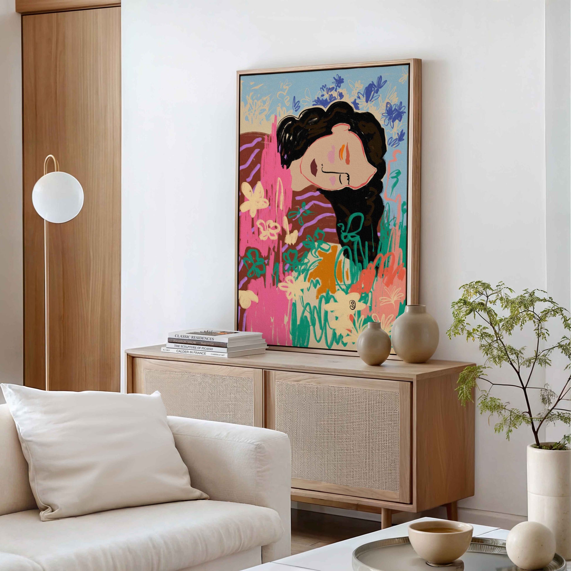 A colorful framed art print featuring a stylized woman with dark hair and a pink face, displayed on a light wood sideboard in a modern living room setting.