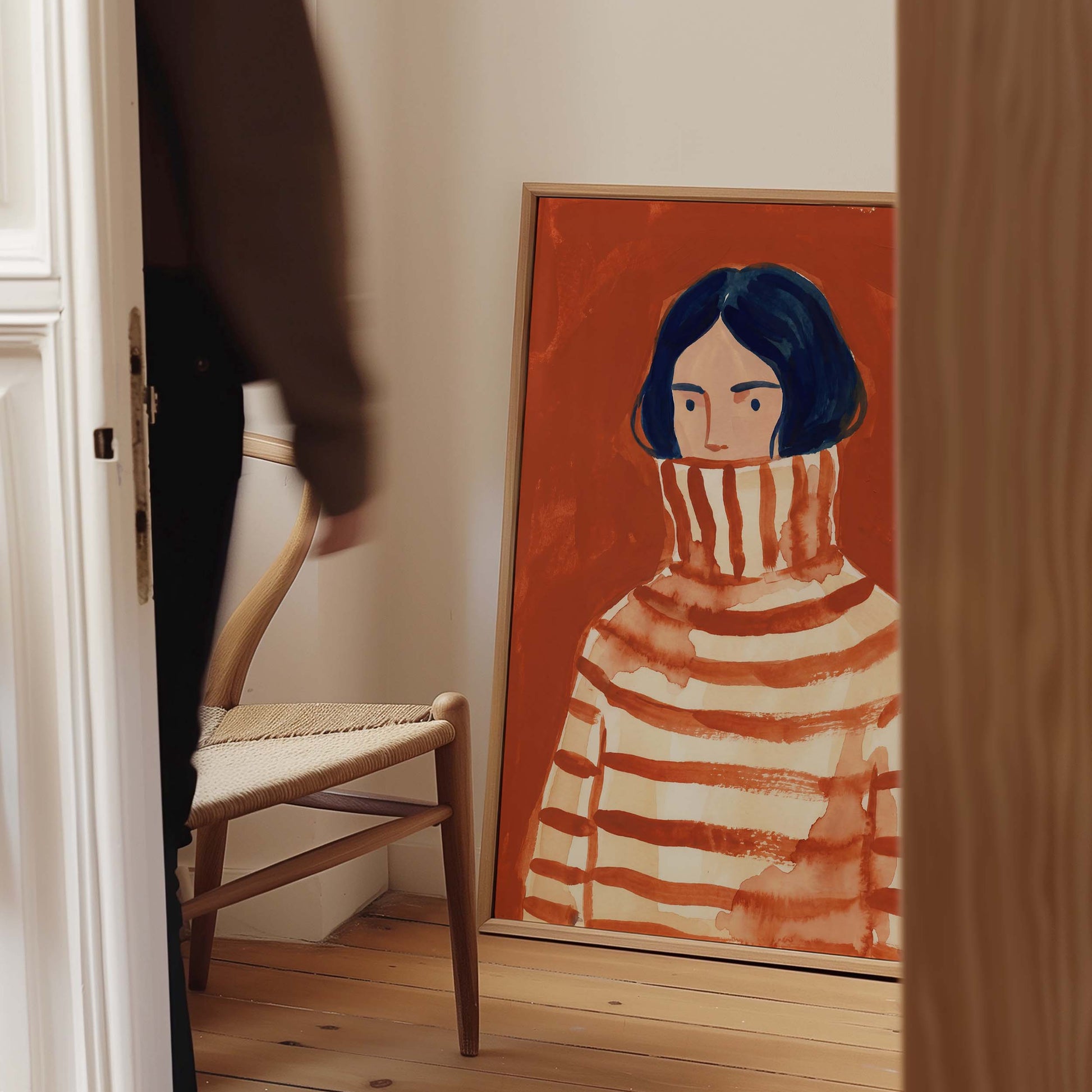 A framed art print depicting a stylized portrait of a person in a striped turtleneck sweater, leaning against a wall in a home interior next to a wooden chair.