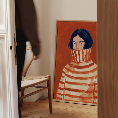 A framed art print depicting a stylized portrait of a person in a striped turtleneck sweater, leaning against a wall in a home interior next to a wooden chair.