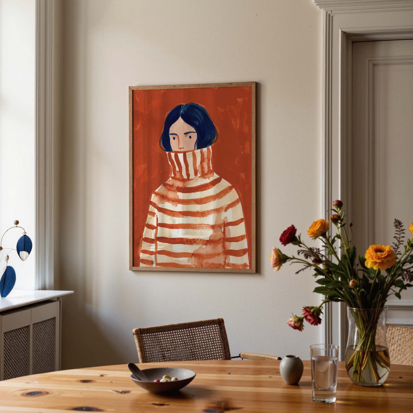 A framed art print depicting a woman in a striped turtleneck sweater hangs on a beige wall in a dining room setting with a wooden table, chairs, and a vase of flowers.