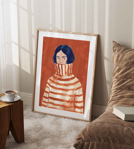 A framed art print leaning against a wall in a cozy room setting. The print features a stylized illustration of a person with dark blue hair wearing a red and white striped turtleneck sweater against a warm orange-red background.