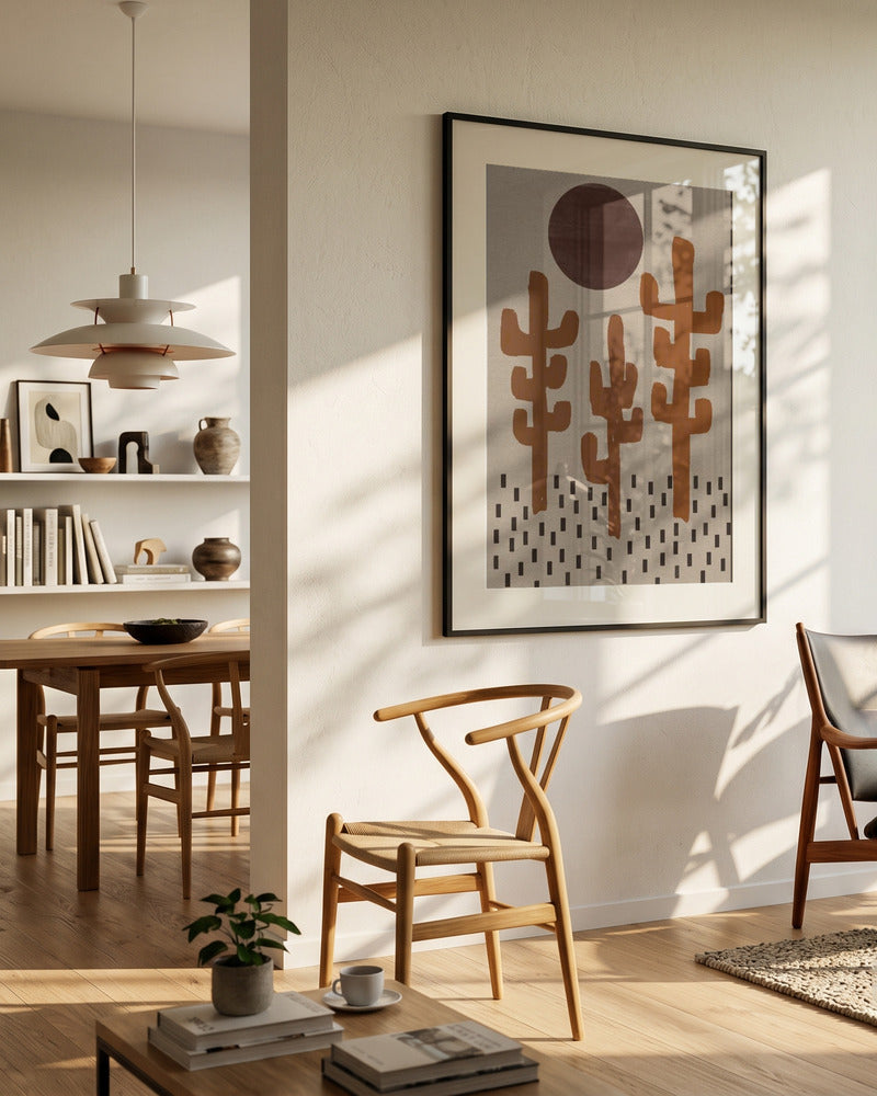 A framed abstract art print featuring stylized cacti hangs on a warm beige wall in a modern, sunlit living space with wooden chairs, a dining table, and a pendant light.