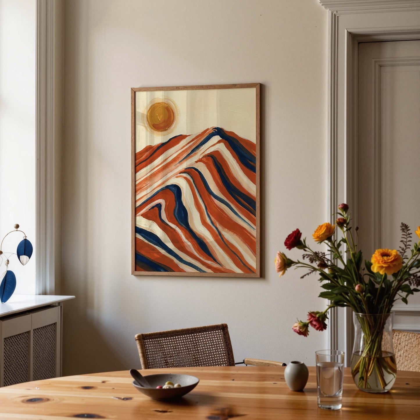 A framed abstract landscape painting with wavy orange, red, cream, and blue mountain-like lines and a yellow sun, displayed on a beige wall above a wooden dining table with flowers and tableware in a real interior setting.