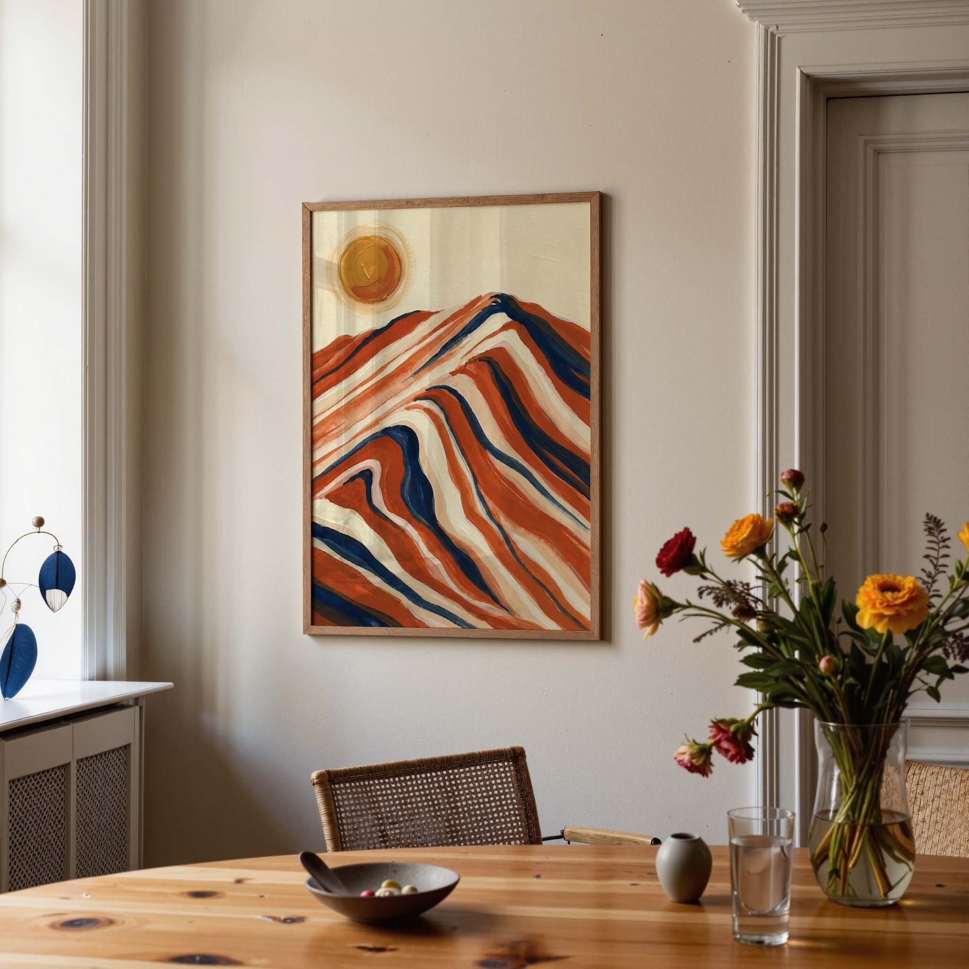A framed abstract landscape painting with wavy orange, red, cream, and blue mountain-like lines and a yellow sun, displayed on a beige wall above a wooden dining table with flowers and tableware in a real interior setting.