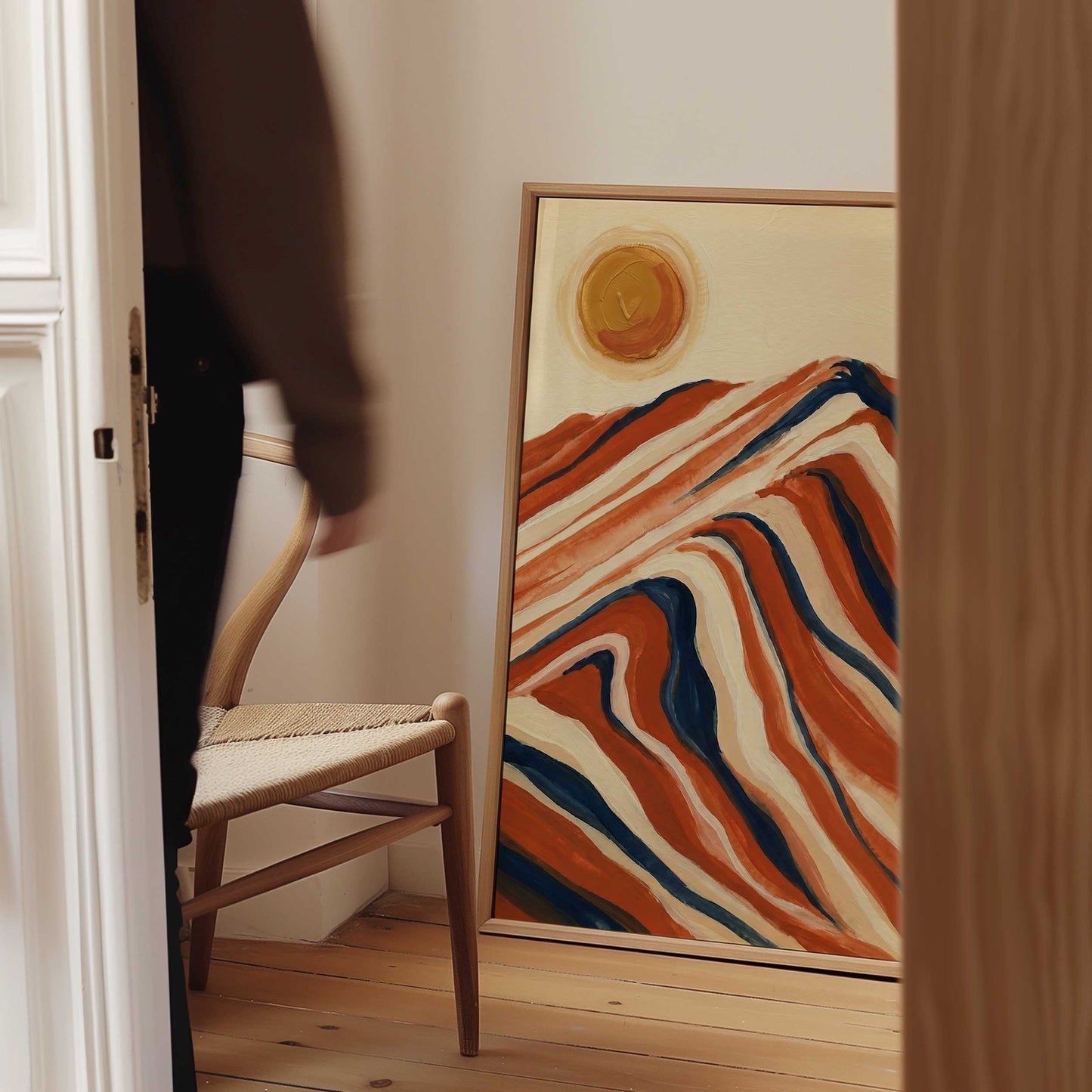 A framed abstract landscape painting with wavy orange, cream, and blue mountain-like forms and a yellow sun, leaning against a wall in a room with a wooden chair and a person partially visible in the doorway.