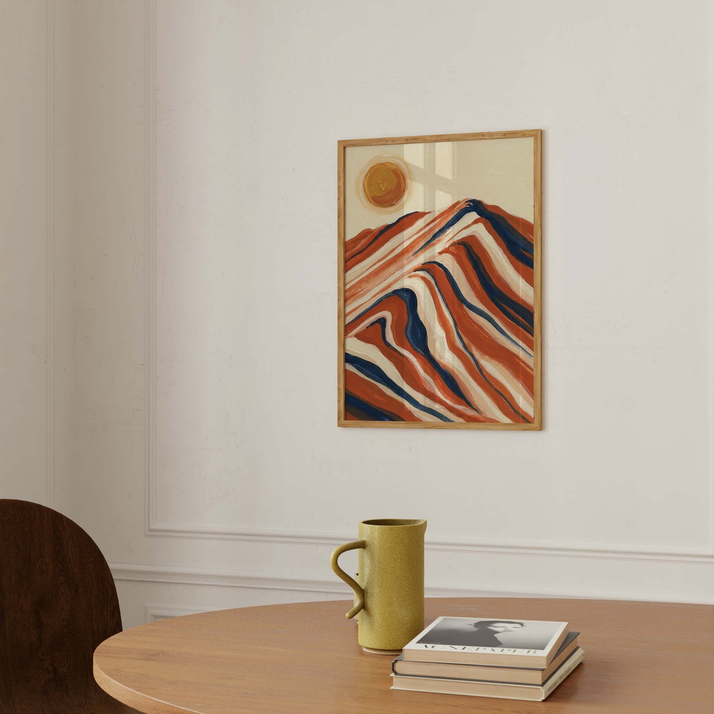 A framed abstract art print featuring wavy red, orange, and blue stripes with a gold sun on a cream background, hanging on a white wall above a wooden table with a green mug and books.