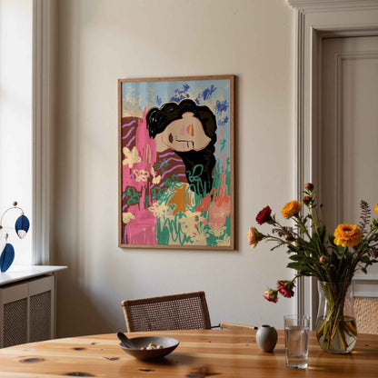 A framed colorful painting of a stylized woman with dark hair hangs on a beige wall above a wooden dining table with flowers and tableware in a real interior setting.