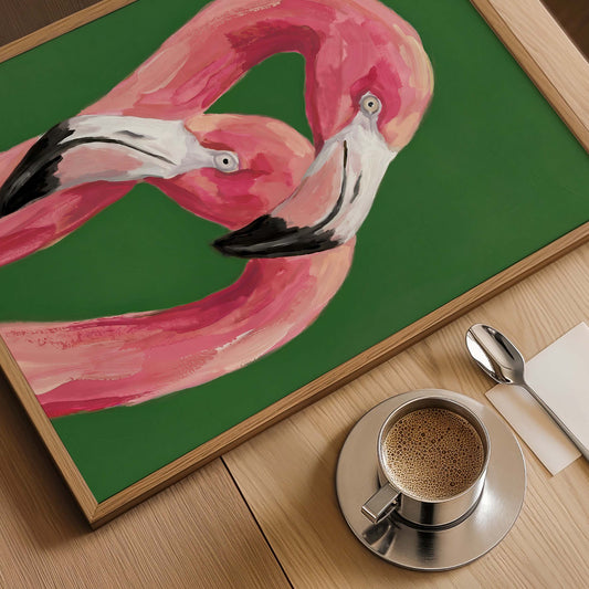 A framed painting of two pink flamingos on a green background, styled on a wooden table next to a cup of coffee, a spoon, and a napkin.