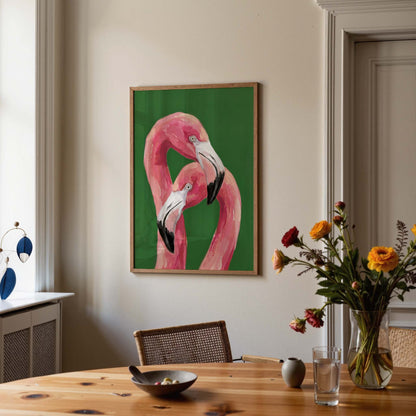 A framed art print of two pink flamingos on a green background, hanging on a beige wall in a styled dining room setting with a wooden table, flowers, and a chair.