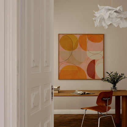 A framed abstract geometric wall art print with overlapping circles in orange, yellow, pink, and brown tones, displayed in a modern room above a wooden desk with a chair and a white cloud-shaped pendant light.