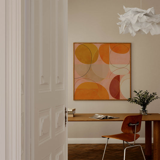A framed abstract geometric wall art print with overlapping circles in orange, yellow, pink, and brown tones, displayed in a modern room above a wooden desk with a chair and a white cloud-shaped pendant light.