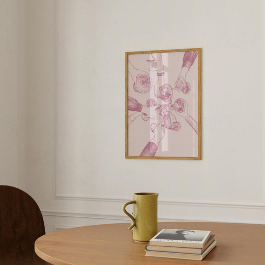 A framed art print featuring a pink and purple botanical illustration with a light wood frame, hanging on a cream wall above a wooden table with a green ceramic mug and a stack of books.