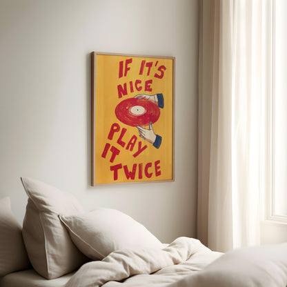 A framed wall art print with the phrase 'IF IT'S NICE PLAY IT TWICE' in red lettering over a yellow background, featuring a red vinyl record illustration, hanging above a bed in a softly lit bedroom.
