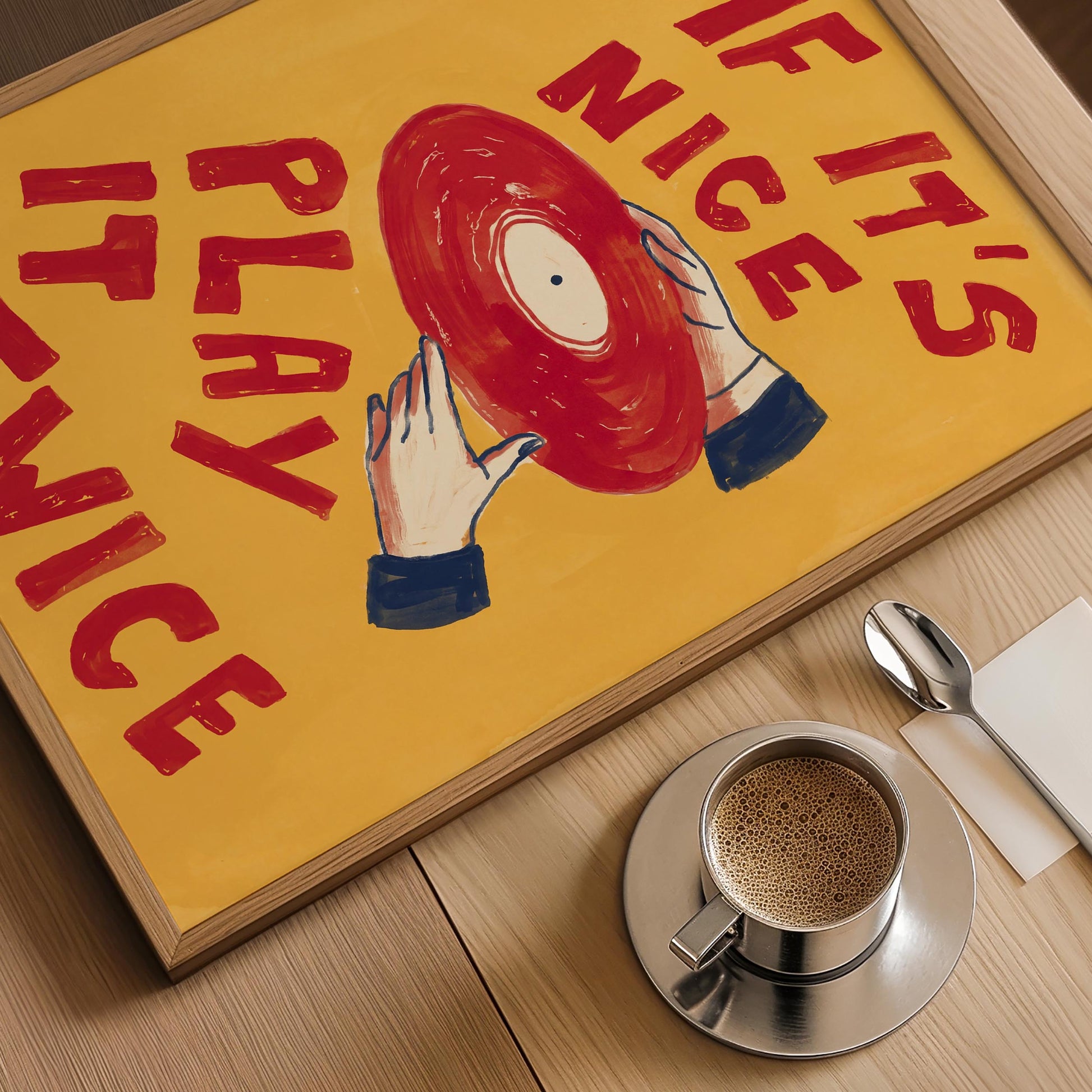 A framed art print featuring a yellow background with red text reading 'IF IT'S NICE PLAY IT TWICE' and an illustration of hands holding a red vinyl record, displayed on a wooden table next to a cup of coffee, a spoon, and a napkin.