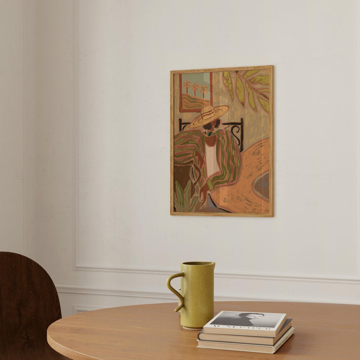 A framed art print featuring an abstract painting of a woman in a hat, hanging on a white wall above a wooden table with a green ceramic mug and a stack of books.