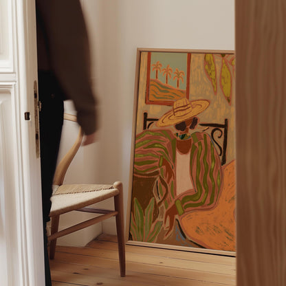 A framed colorful painting leaning against a white wall in a room, showing a seated woman in a wide-brim straw hat and sunglasses with palm trees and abstract foliage. A blurred person walks past a chair on the left, and a wooden door frame borders the scene.