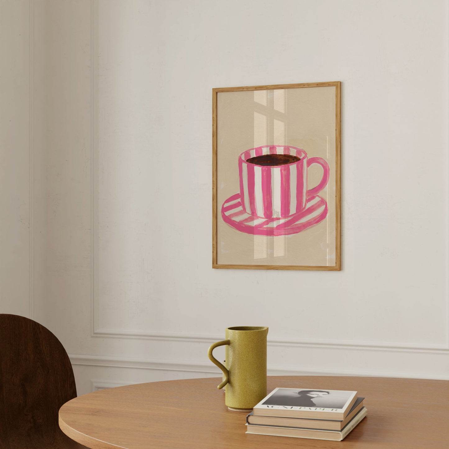 A framed art print featuring a pink and white striped coffee cup and saucer, displayed on a white wall above a wooden table with a green ceramic mug and a stack of books.