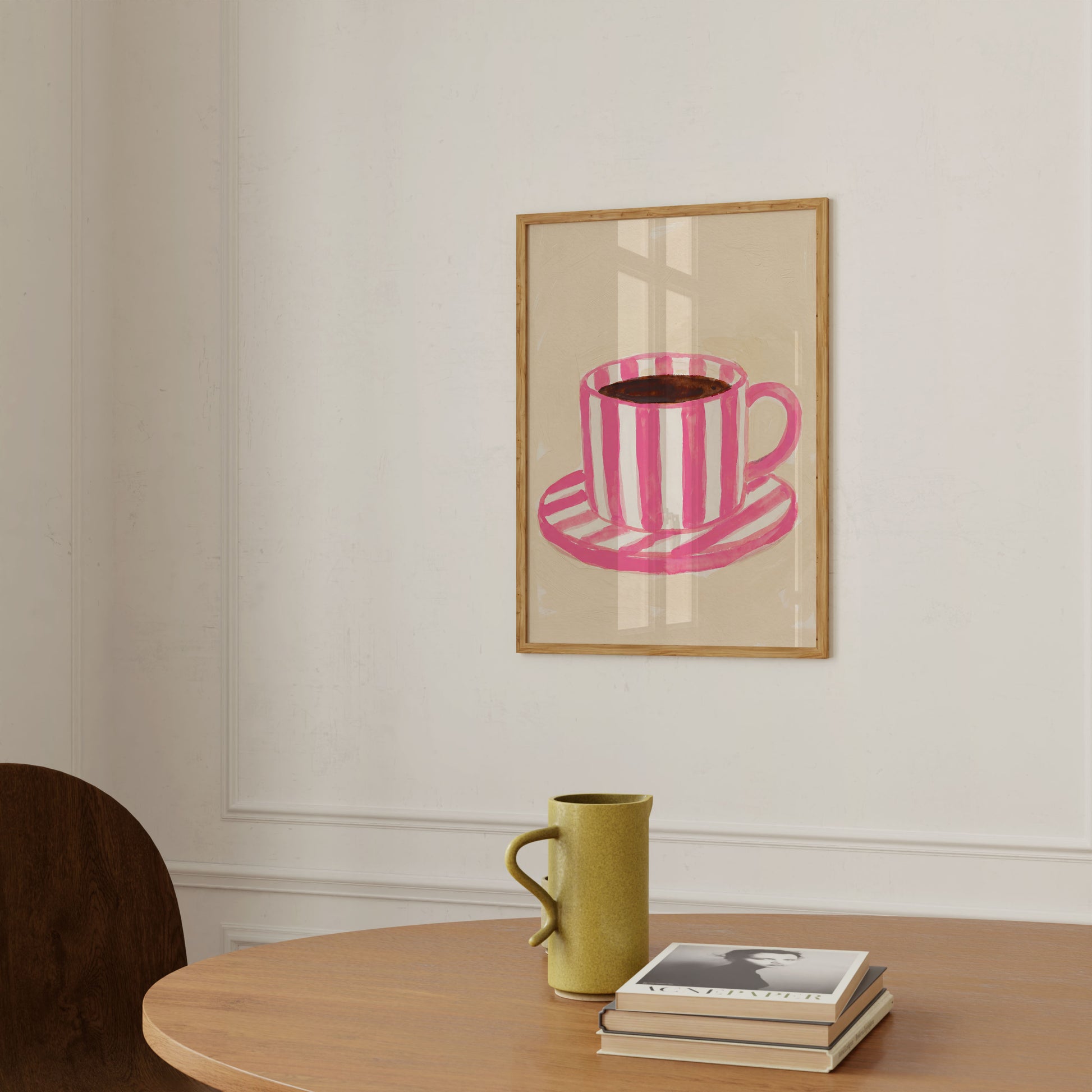 A framed art print featuring a pink and white striped coffee cup and saucer, displayed on a white wall above a wooden table with a green ceramic mug and a stack of books.