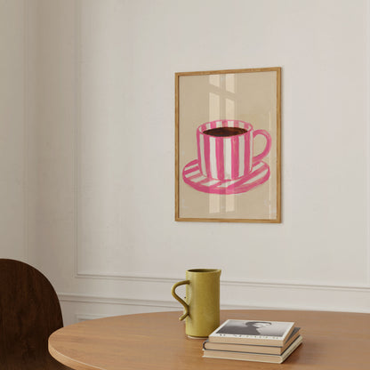 A framed art print featuring a pink and white striped coffee cup and saucer, displayed on a white wall above a wooden table with a green ceramic mug and a stack of books.